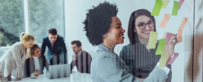 Two smiling women collaborate on a glass wall covered in colorful sticky notes in a bright conference room, while three colleagues work together at a table in the background.