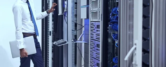 A male IT professional in a white dress shirt and blue tie holds a laptop while operating a monitor in a server room filled with rack-mounted equipment and blue cables.
