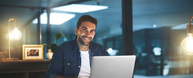 A smiling man in a denim shirt sits at a warmly lit wooden desk with an open laptop, notebook, and small succulent in a moody office setting.