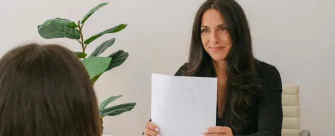 A woman with long dark hair in a black blazer holds printed documents across a white desk from another person in a bright, minimalist office.