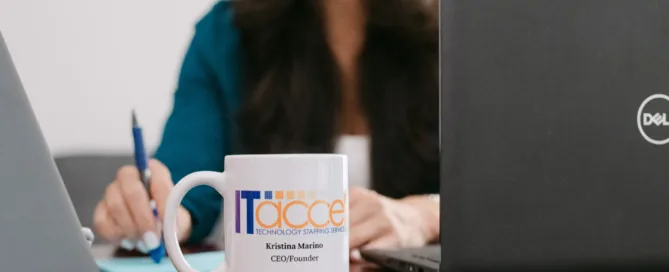An ITaccel branded mug for Kristina Marino, CEO/Founder, in the foreground on a dark wood desk with a smiling woman in a teal blazer writing notes between two laptops in the background.