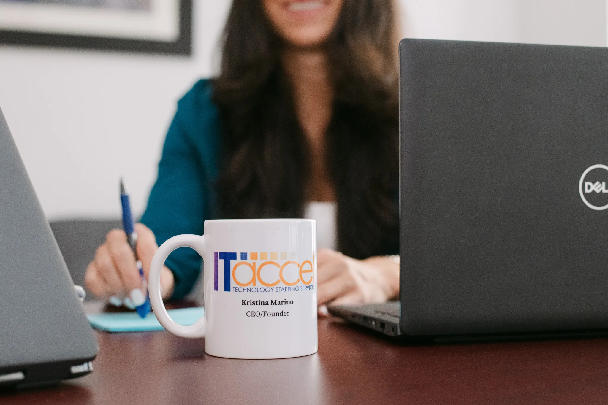 Close-up of an IT Accel coffee mug on an office desk with a woman working on a laptop in the background.