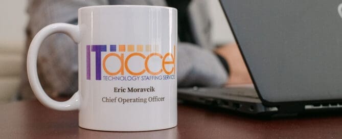A white ITaccel branded mug for Eric Moravcik, Chief Operating Officer, sitting on a dark wood desk beside an open Dell laptop.