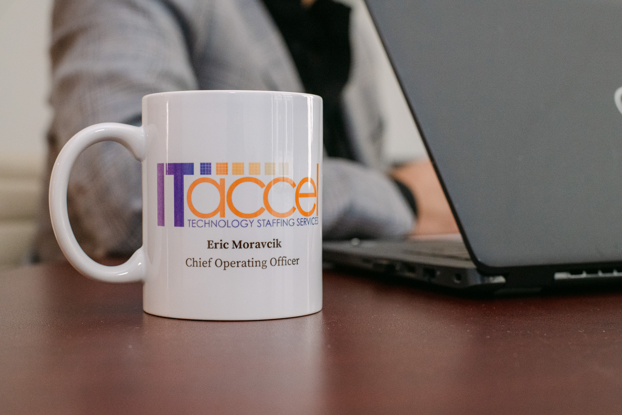 IT Accel coffee mug with company logo and COO name on a desk beside a laptop.