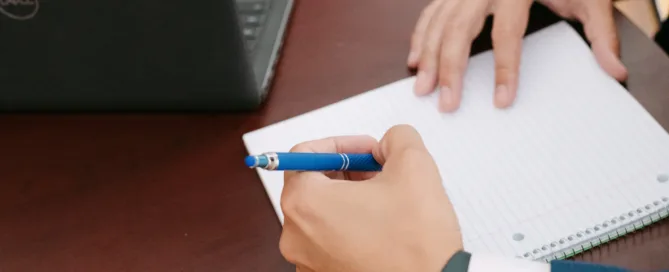 A close-up of two people's hands over an open spiral notebook on a dark wood desk, with a Dell laptop visible in the background.