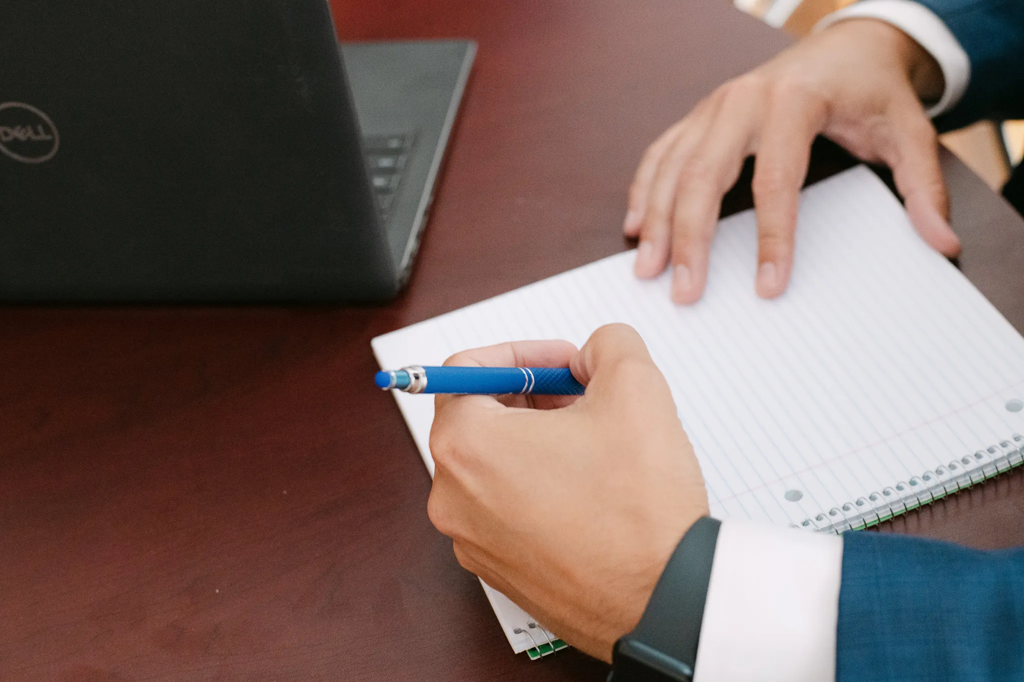 Close-up of a business professional writing notes in a notebook beside a laptop.