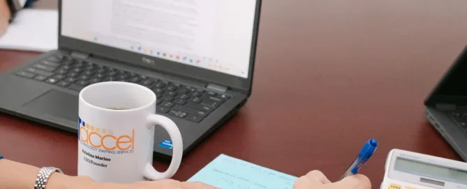 A close-up of a woman's hands writing on a blue notepad beside an ITaccel branded mug, laptop, and calculator on a dark wood desk.