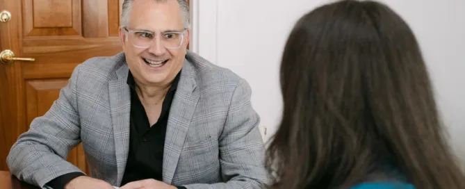 A smiling man in a gray plaid blazer and clear-framed glasses sits across a desk from a woman in a teal top during a professional meeting.