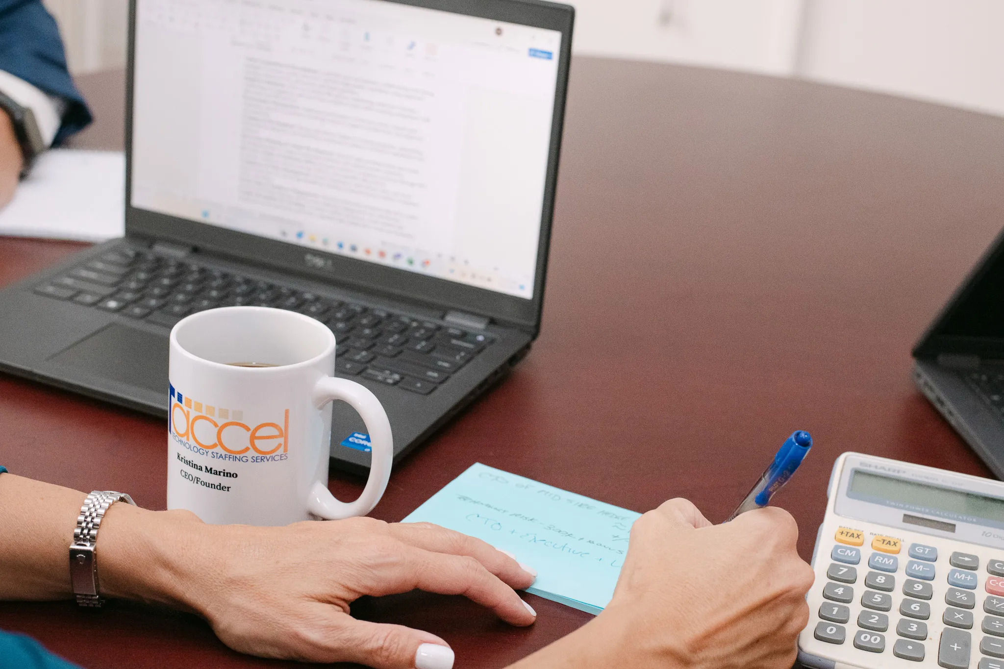 IT Accel team member reviewing notes and staffing data at a desk with a laptop, calculator, and branded mug during an IT recruitment planning session.