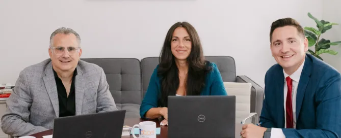 Three smiling ITaccel team members seated at a dark wood conference table with Dell laptops and branded mugs in a modern office.