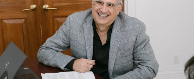 A smiling man in a gray plaid blazer and clear-framed glasses sits at a dark wood desk with a Dell laptop, calculator, and documents in a traditional office setting.