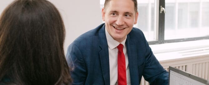 A smiling man in a blue suit and red tie engaged in a conversation with a woman across a desk in a bright office.