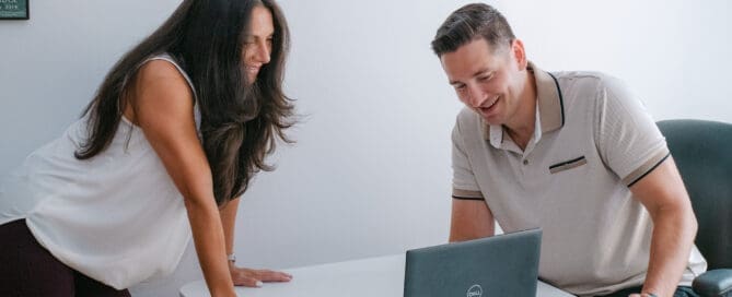 Two colleagues smiling and leaning over a white desk to view a Dell laptop in a bright office setting.