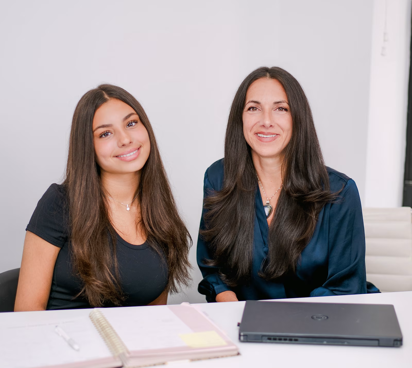 two-team-members-at-desk-it-accel. Two smiling IT Accel team members seated at a white desk with a spiral planner and a closed laptop