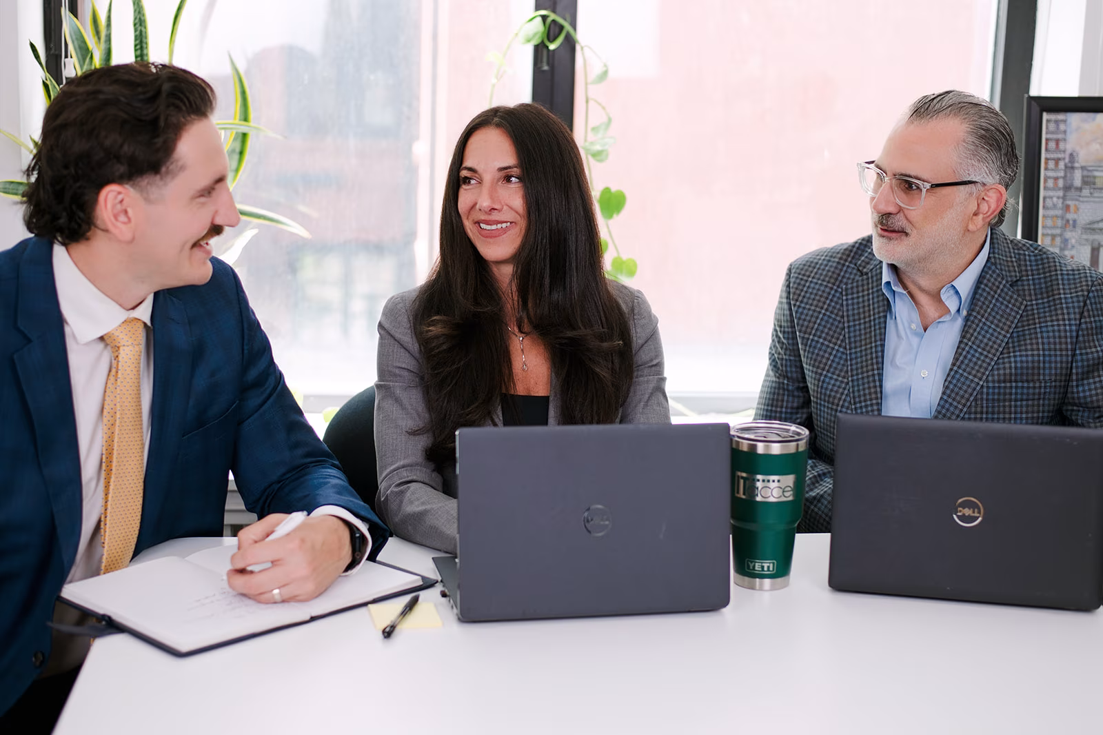 team-collaborating-in-office-meeting-it-accel. Three IT Accel team members talking and smiling during a meeting at a desk with laptops and an open notebook