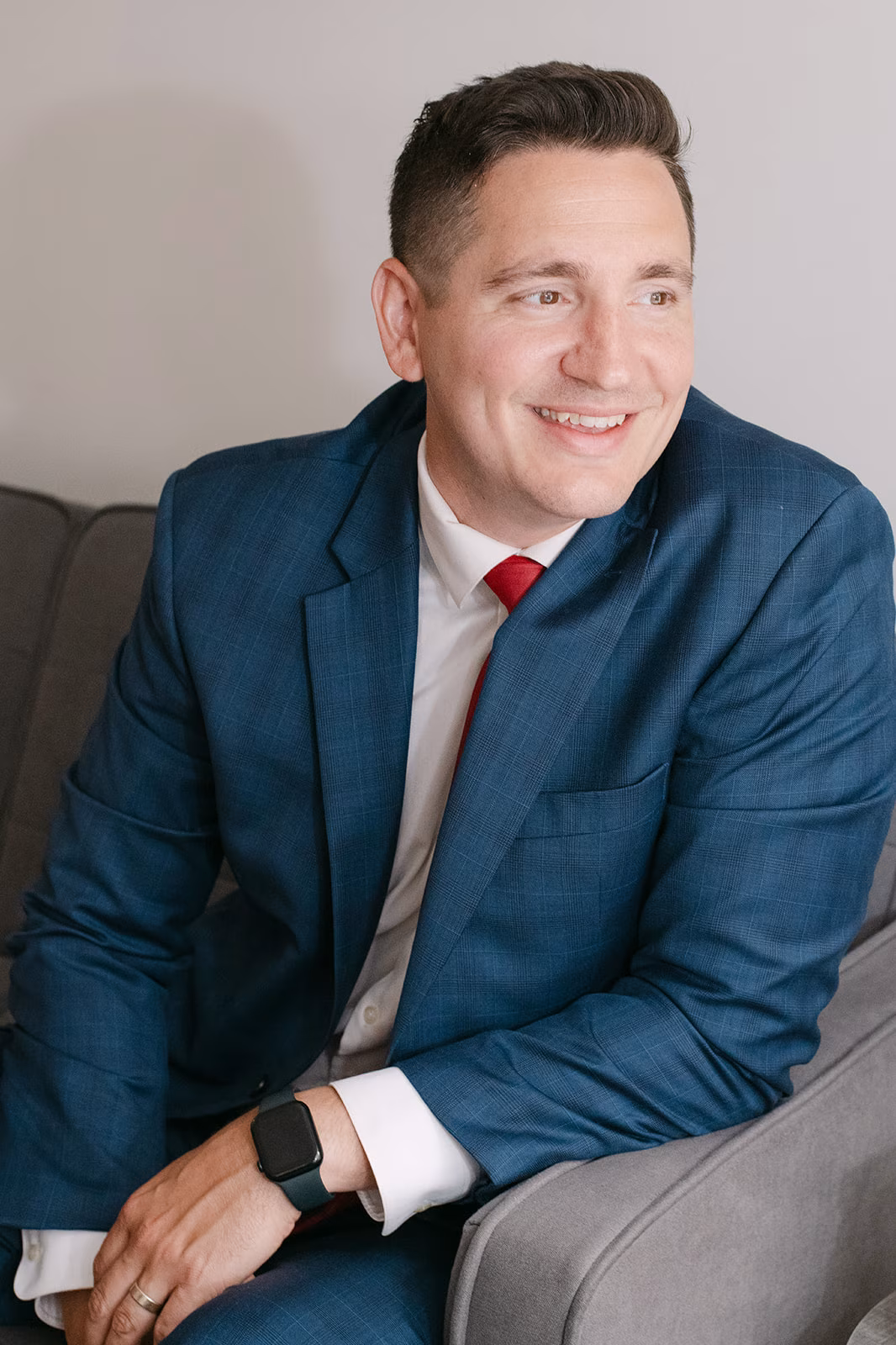 professional-portrait-in-navy-suit-it-accel. Smiling IT Accel team member in a navy plaid suit and red tie, seated on a gray sofa and looking off to the side